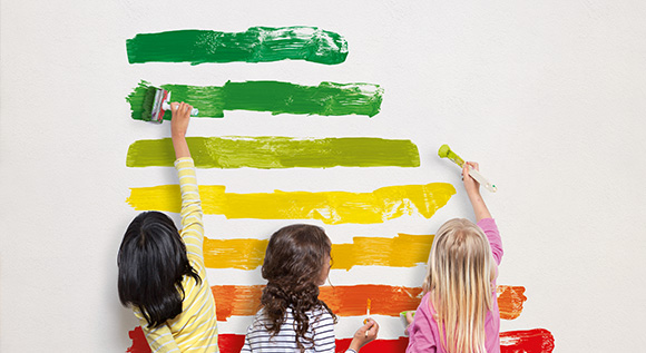 Drei Kinder malen die Effizienzstufen des EU-Energielabels mit Pinseln an eine Wand.