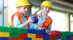 Zwei Jungs mit Bauhelmen spielen mit großen Legosteinen.