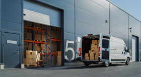 Ein Transporter voller Kartons vor einer Lagerhalle mit weiteren Kartons.